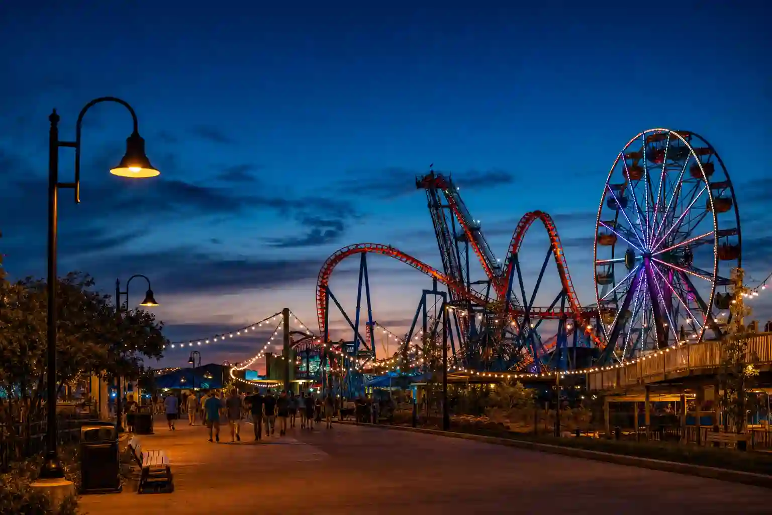 Amusement park midway at dusk with a lit roller coaster and Ferris wheel