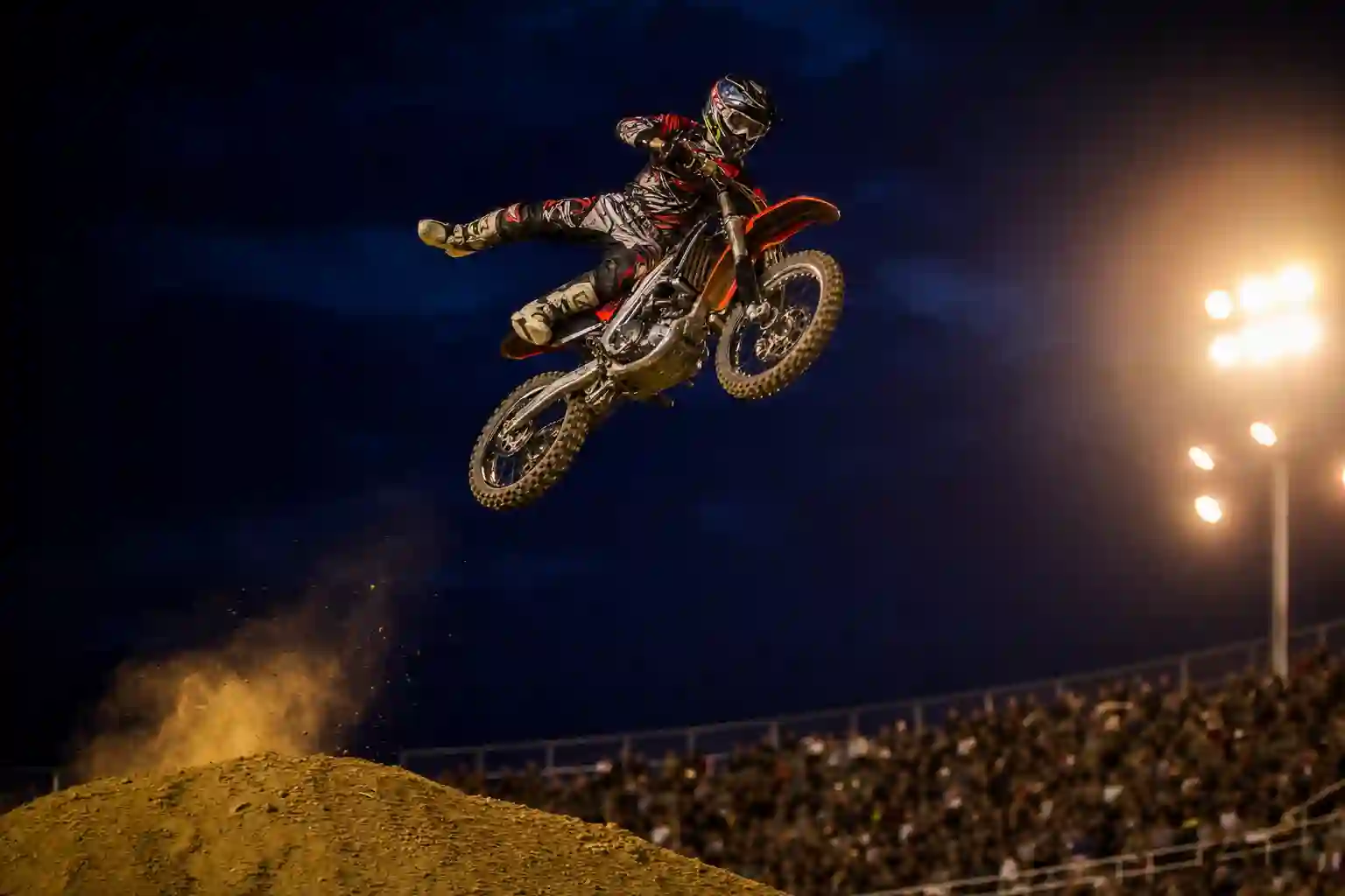 A motocross stunt rider mid-air over a dirt jump at a fairground arena